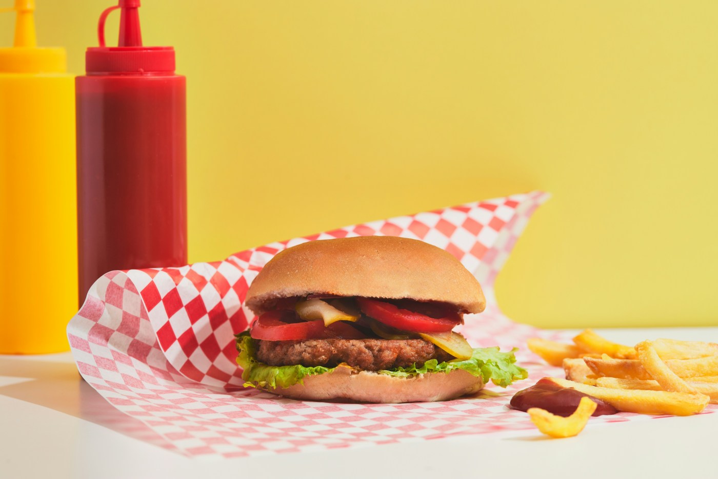 Cheap burger with ketchup on red and white chequered paper tablecloth on bright yellow background symbolising fast food
